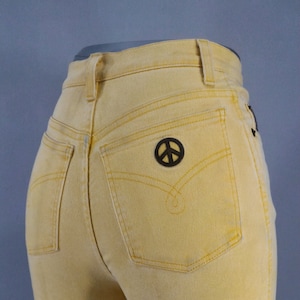 May include: Yellow denim jeans with a high waist and a peace sign patch on the back pocket. The jeans have yellow stitching and a classic five-pocket design. The overall style is vintage.