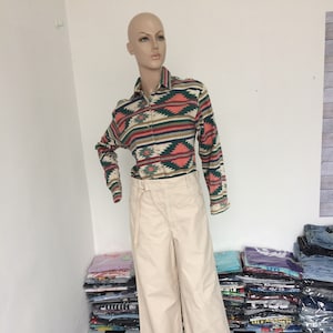 Puede incluir: Una camisa blanca de manga larga con un patrón geométrico en tonos de verde, rosa y marrón. La camisa se lleva con un pantalón de pierna ancha de color crema.