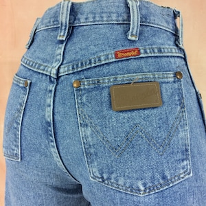May include: A pair of light blue denim jeans with a brown leather Wrangler label on the back pocket. The jeans have a high waist and a classic straight leg fit.