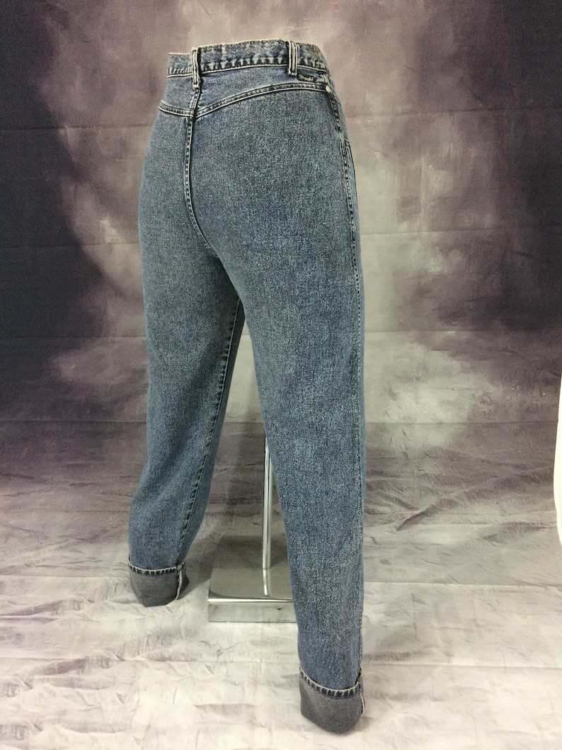 May include: Acid wash denim jeans with a high waist and cuffed hems. The jeans have a classic five-pocket design and a button and zipper closure. The denim has a faded, textured appearance.