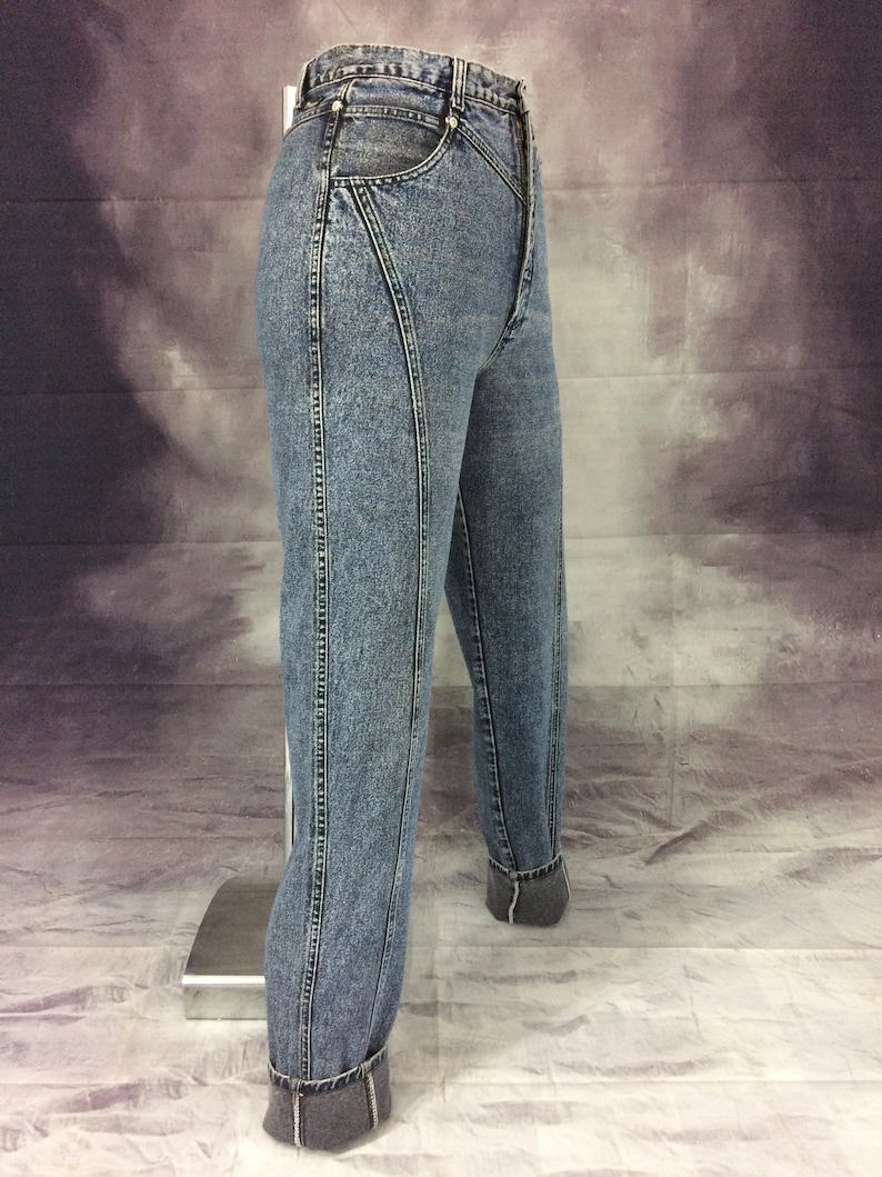May include: Light blue denim jeans with a high waist and a tapered leg. The jeans feature a unique seam design on the front and back, with cuffed hems. The denim has a faded, acid-wash appearance, and the jeans are displayed on a mannequin.