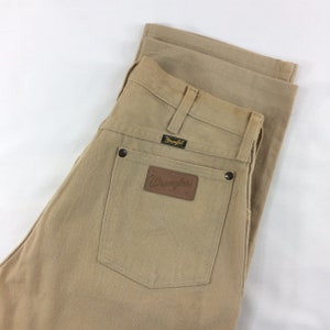 May include: A pair of tan Wrangler jeans with a brown leather patch on the back pocket. The jeans are folded in half and the back pockets are visible.