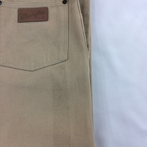 May include: A pair of tan Wrangler jeans with a brown leather patch on the back pocket. The patch has the Wrangler logo in white.