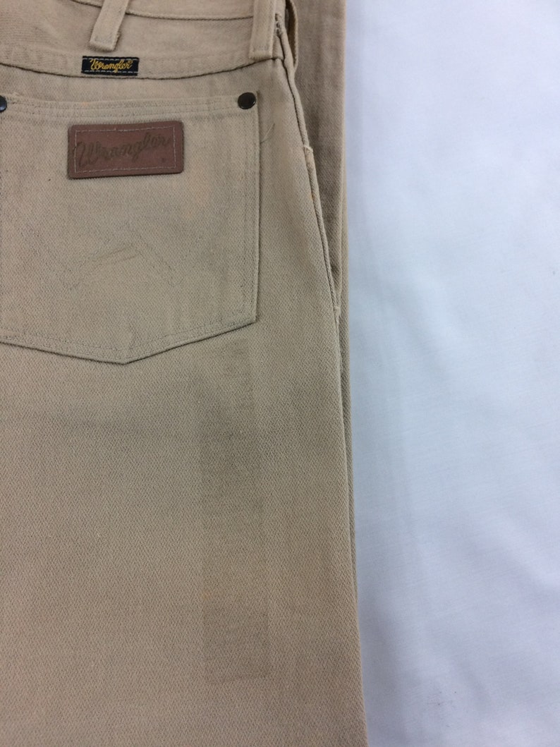 May include: A pair of tan Wrangler jeans with a brown leather patch on the back pocket. The patch has the Wrangler logo in white.