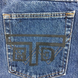 May include: A close-up of a blue denim pocket with a black graphic design of a stylized letter 'T' with two squares inside. The pocket has brown stitching around the edges.