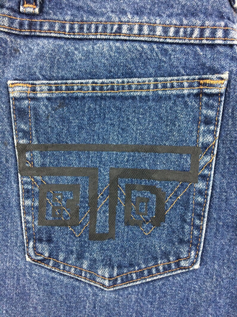 May include: A close-up of a blue denim pocket with a black graphic design of a stylized letter 'T' with two squares inside. The pocket has brown stitching around the edges.