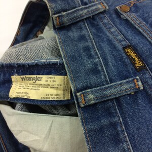 Peut inclure: Un gros plan sur une paire de jeans en denim bleu avec une &eacute;tiquette Wrangler. L'&eacute;tiquette indique la taille 31 x 34 et le texte "Machine wash hot and inside out first three times with like colors, tumble dry remove promptly. Made in USA 100% Cotton. C478-003 051071 501 750."
