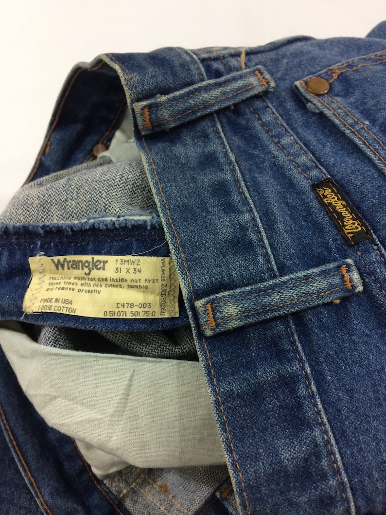Peut inclure: Un gros plan sur une paire de jeans en denim bleu avec une &eacute;tiquette Wrangler. L'&eacute;tiquette indique la taille 31 x 34 et le texte "Machine wash hot and inside out first three times with like colors, tumble dry remove promptly. Made in USA 100% Cotton. C478-003 051071 501 750."