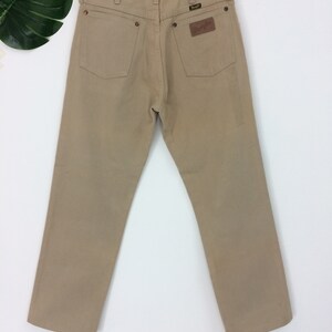 May include: A pair of tan-colored straight-leg jeans with a brown leather patch on the back pocket that says "Wrangler".