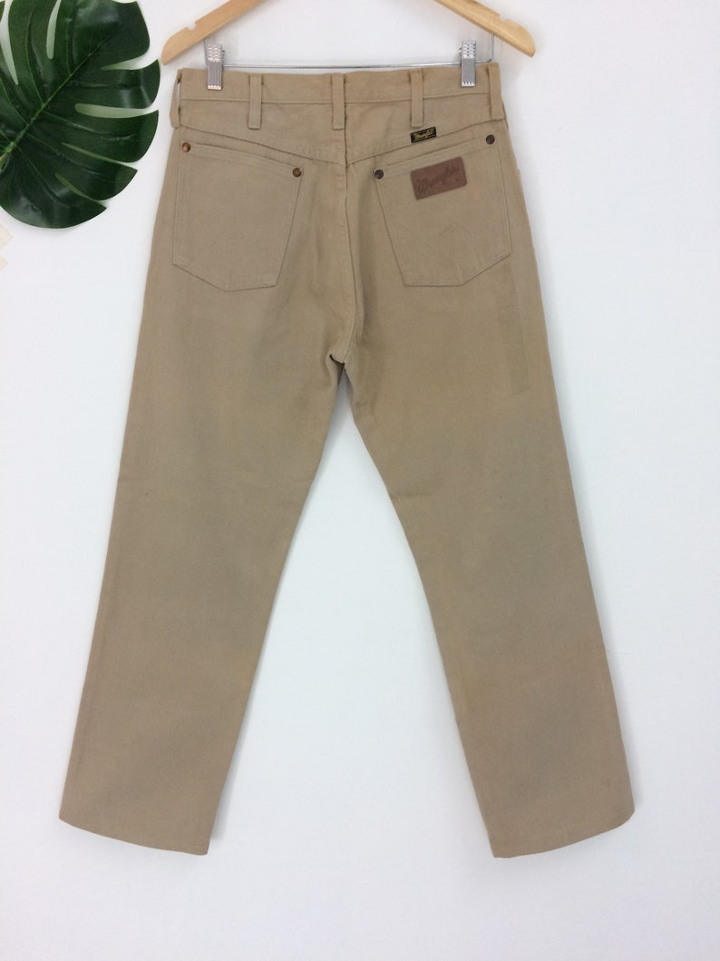 May include: A pair of tan-colored straight-leg jeans with a brown leather patch on the back pocket that says "Wrangler".
