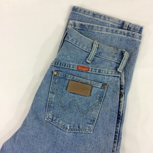 May include: A pair of light blue denim jeans with a brown leather patch on the back pocket. The patch has the word "Wrangler" printed on it in red.