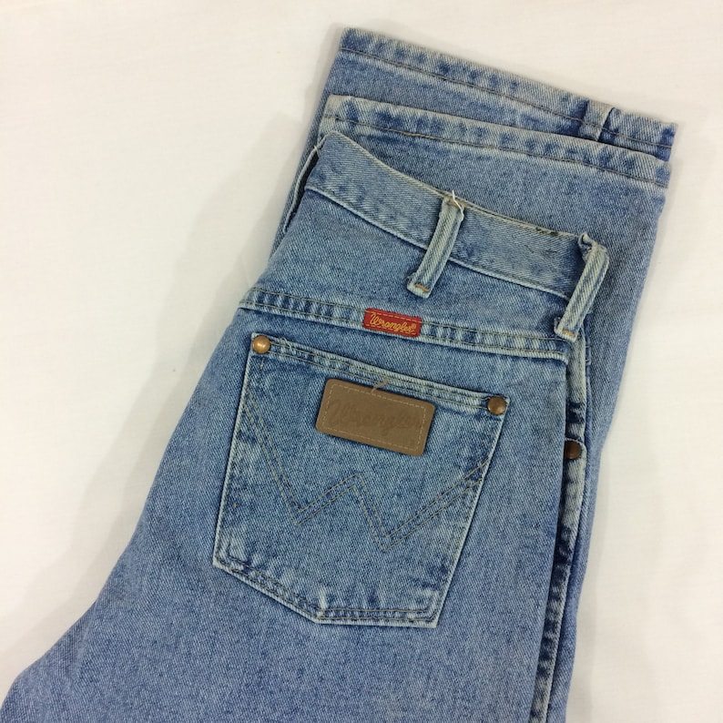 May include: A pair of light blue denim jeans with a brown leather patch on the back pocket. The patch has the word "Wrangler" printed on it in red.