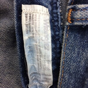 May include: Close-up of a blue denim jean with a white tag sewn into the seam. The tag has text on it, but it is too blurry to read.