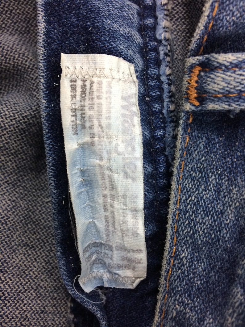 May include: Close-up of a blue denim jean with a white tag sewn into the seam. The tag has text on it, but it is too blurry to read.