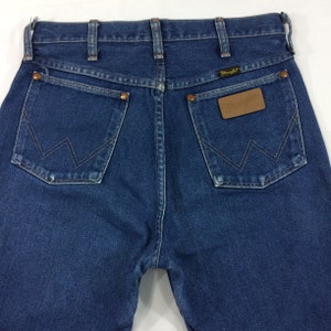 May include: Close-up of the back of blue denim jeans. The jeans have two back pockets with a signature "W" stitch design. A brown leather patch with the "Wrangler" logo is on the right pocket. The jeans have copper rivets and a classic five-pocket design.