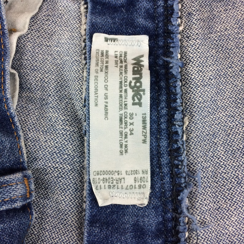 May include: A white care label attached to a pair of blue denim jeans. The label reads "Wrangler 13MWZPW 30 X 34", "Machine wash cold with like colors. Only non-chlorine bleach when needed, tumble dry low or line dry", "Made in Mexico of US fabric, 100% cotton, exclusive of decoration", "RN 130273 1500000260", "70916 LAR-E049-019", "081071128117".