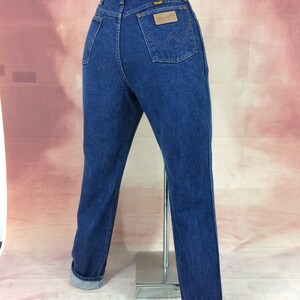 May include: Dark blue denim jeans with a classic fit, featuring a Wrangler label on the back pocket. The jeans have a straight leg and are cuffed at the ankles. The jeans are displayed on a mannequin.