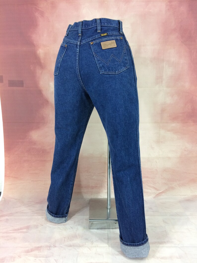 May include: Dark blue denim jeans with a classic fit, featuring a Wrangler label on the back pocket. The jeans have a straight leg and are cuffed at the ankles. The jeans are displayed on a mannequin.