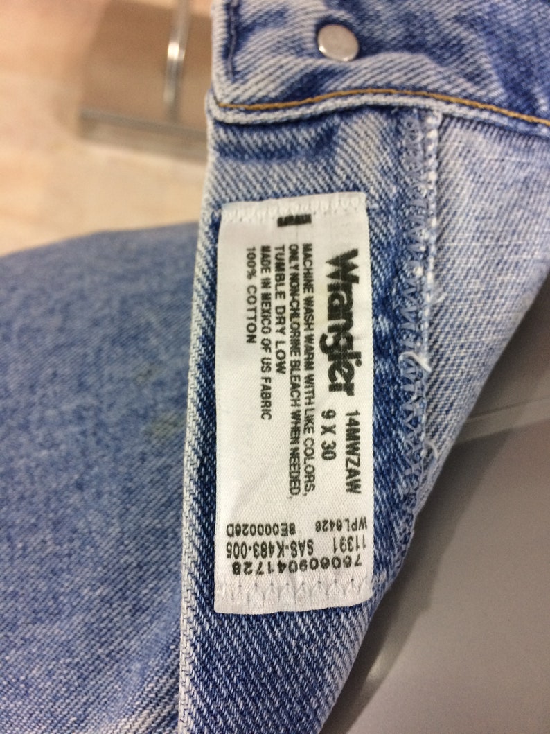 May include: A white Wrangler jeans care label with black text. The label says "Machine wash warm with like colors, only non-chlorine bleach when needed. Tumble dry low. Made in Mexico of US fabric. 100% cotton. Wrangler 14MWZAW 9 X 30"