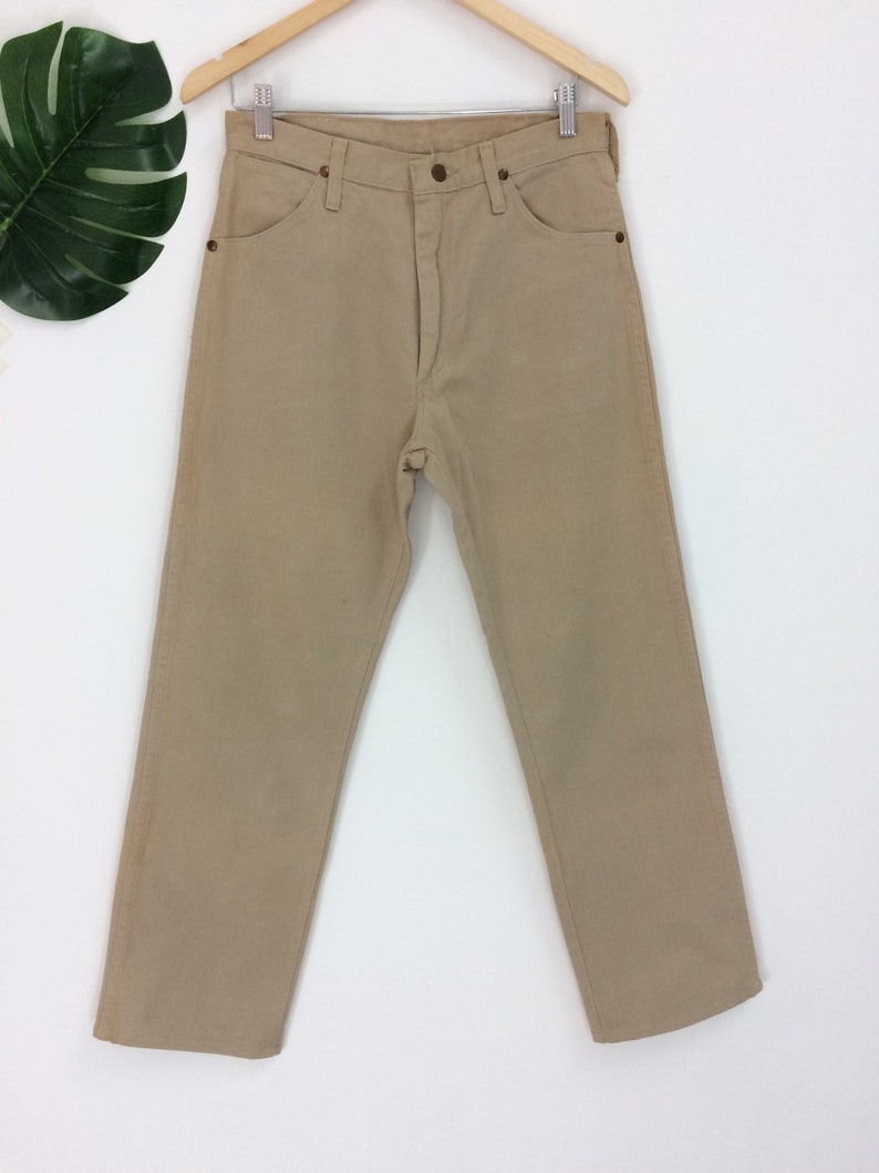 May include: A pair of light brown straight leg jeans hanging on a wooden hanger against a white background.