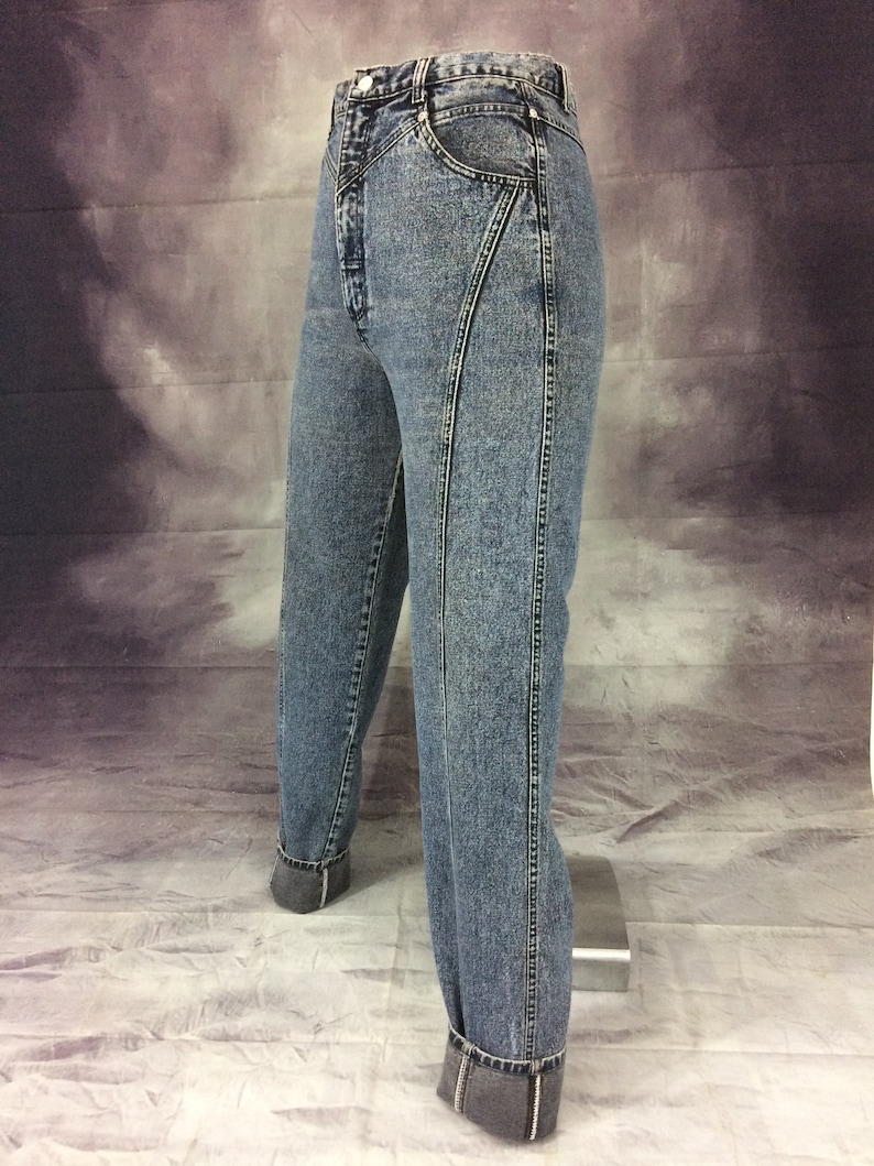May include: Acid wash denim jeans with a high waist and unique seam detailing. The jeans have a classic five-pocket design and are cuffed at the ankles. The light blue wash and tailored fit give a vintage aesthetic.