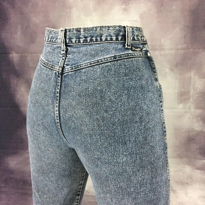 May include: Close-up of light blue denim jeans. The jeans have a high waist and a classic design. The back of the jeans is visible, showing the stitching and the Wrangler label.
