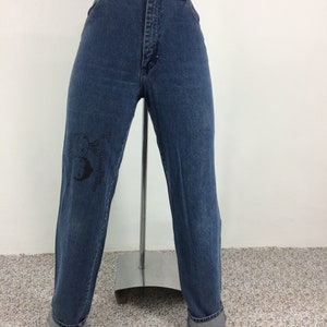 May include: A pair of blue denim jeans with a black graphic on the left leg. The jeans have a high waist and a straight leg fit.