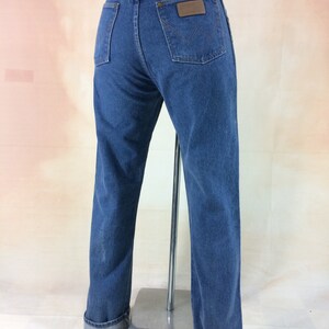 Peut inclure: Un jean bleu denim avec un &eacute;cusson en cuir marron sur la poche arri&egrave;re qui dit "Wrangler". Le jean a une jambe droite et est retrouss&eacute; en bas.
