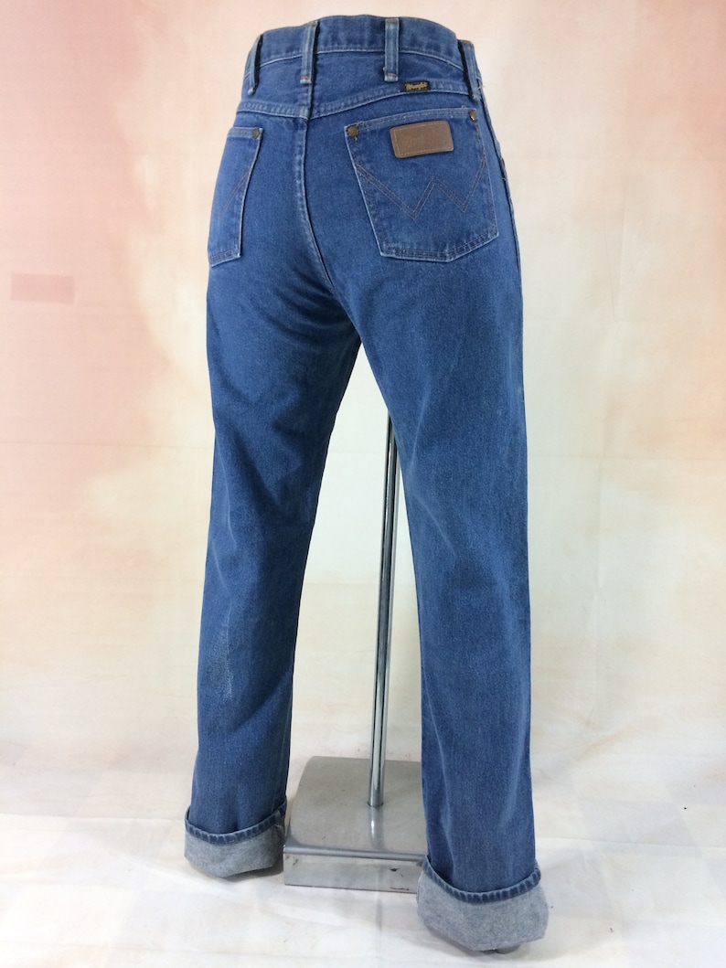 Peut inclure: Un jean bleu denim avec un &eacute;cusson en cuir marron sur la poche arri&egrave;re qui dit "Wrangler". Le jean a une jambe droite et est retrouss&eacute; en bas.