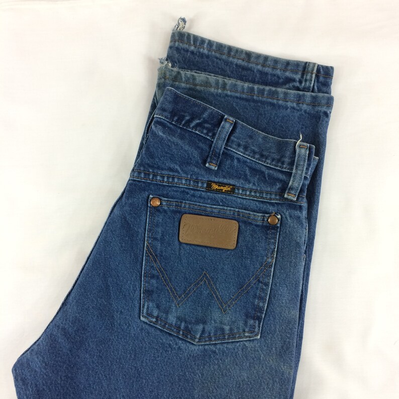 May include: A pair of blue denim jeans with a brown leather patch on the back pocket. The patch has the word "Wrangler" printed on it in gold lettering.
