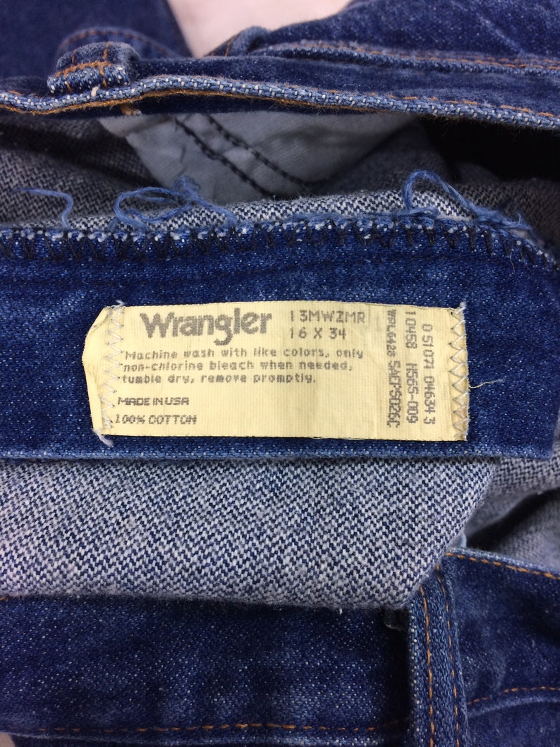 May include: Close-up of blue denim jeans with a Wrangler label. The label indicates the size is 16 x 34 and the material is 100% cotton. The jeans have a classic design and are made in the USA.