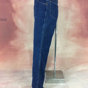 May include: Dark blue denim jeans with a classic straight leg fit. The jeans feature a traditional five-pocket design, a button and zipper closure, and a folded cuff at the hem. The jeans are displayed on a silver metal stand.