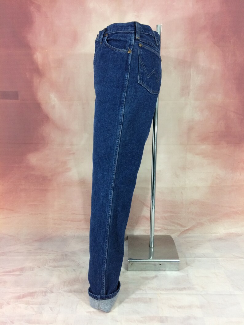 May include: Dark blue denim jeans with a classic straight leg fit. The jeans feature a traditional five-pocket design, a button and zipper closure, and a folded cuff at the hem. The jeans are displayed on a silver metal stand.
