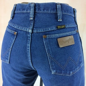 May include: Close-up of the back of blue denim Wrangler jeans. The jeans have two back pockets with copper rivets, a brown leather patch with the Wrangler logo, and a small black tag with the Wrangler logo above the right pocket.