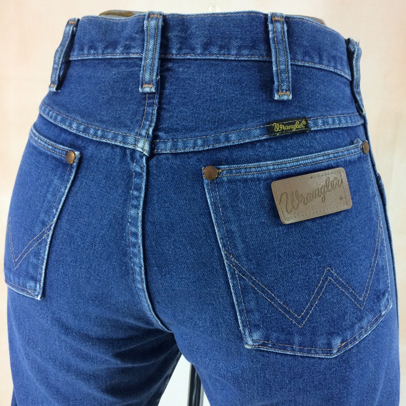 May include: Close-up of the back of blue denim Wrangler jeans. The jeans have two back pockets with copper rivets, a brown leather patch with the Wrangler logo, and a small black tag with the Wrangler logo above the right pocket.