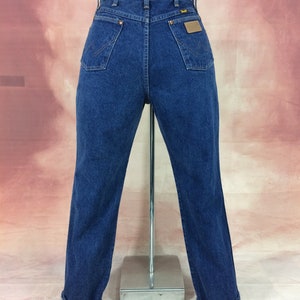 May include: Dark blue denim jeans with a classic fit, featuring a Wrangler brand patch on the back pocket. The jeans have a high waist, two back pockets, and a straight leg with cuffed hems. The jeans are displayed on a mannequin.