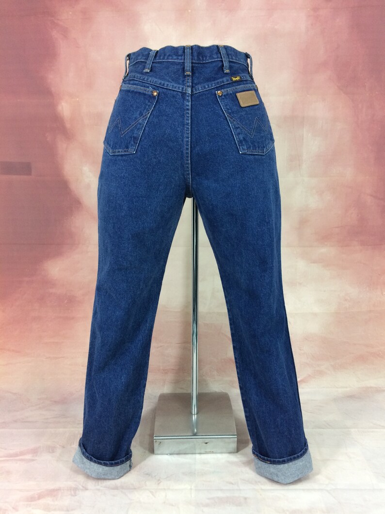 May include: Dark blue denim jeans with a classic fit, featuring a Wrangler brand patch on the back pocket. The jeans have a high waist, two back pockets, and a straight leg with cuffed hems. The jeans are displayed on a mannequin.