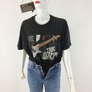 May include: Black Eric Clapton World Tour t-shirt with a graphic of a guitar and the text "LIVE" and "ERIC CLAPTON WORLD TOUR". The shirt is paired with light blue denim jeans. The mannequin is wearing the shirt and jeans.