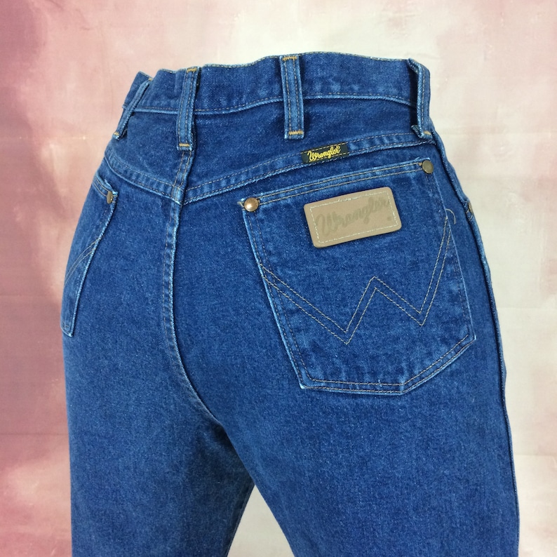 May include: Dark blue denim jeans with a high waist and a classic five-pocket design. The back features a leather patch with the "Wrangler" logo and a signature "W" stitch on the back pocket. The jeans have a straight leg cut.
