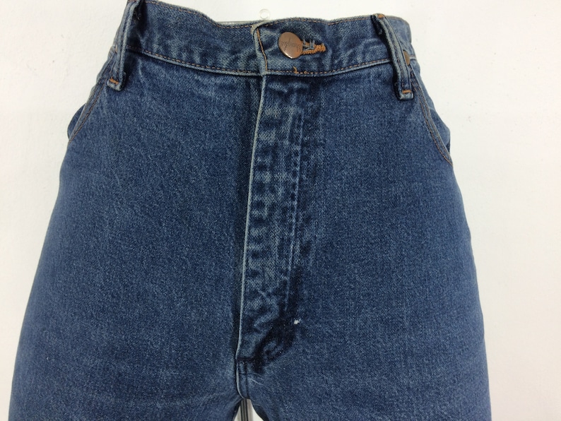 May include: Close-up of the front of a pair of blue denim jeans. The jeans have a button fly and a copper-colored button. The fabric is a medium-wash denim.