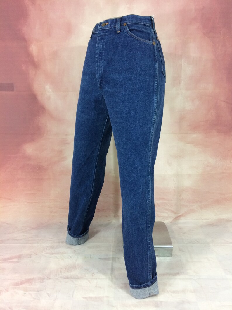 May include: Dark blue denim jeans with a high waist and straight leg. The jeans have a classic five-pocket design, a button and zipper closure, and are cuffed at the ankles. The jeans are displayed on a mannequin.