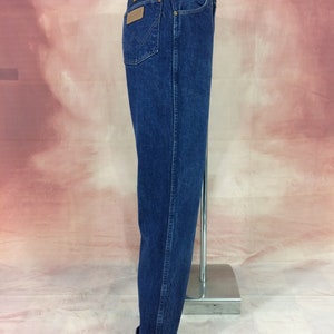 May include: Dark blue denim jeans with a classic fit. Features include a brown leather patch on the back pocket and a straight leg. The jeans are cuffed at the bottom, showcasing a lighter denim lining.