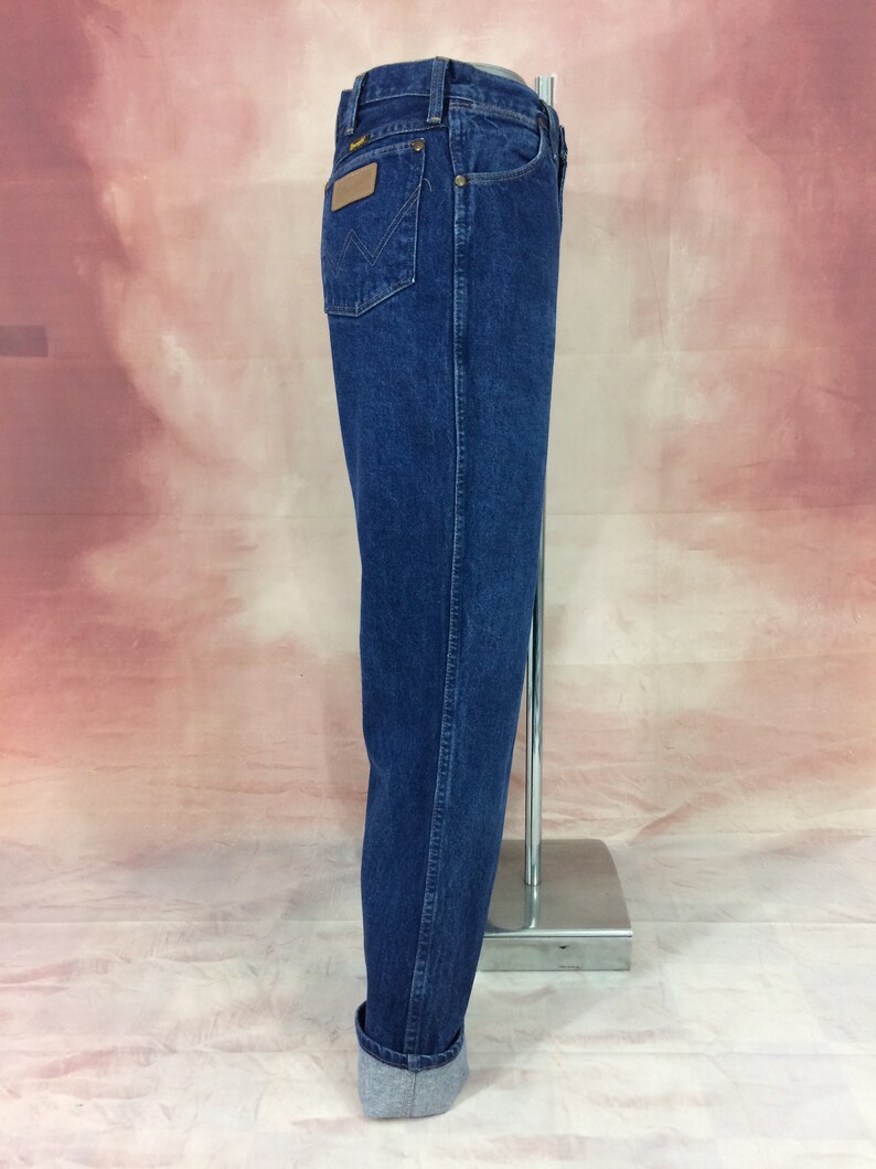 May include: Dark blue denim jeans with a classic fit. Features include a brown leather patch on the back pocket and a straight leg. The jeans are cuffed at the bottom, showcasing a lighter denim lining.