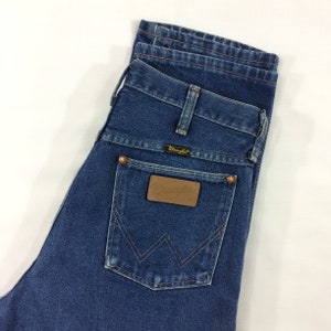 May include: Close-up of blue denim jeans with the Wrangler logo on the back pocket. The jeans have a classic five-pocket design with copper-colored rivets and a leather patch.