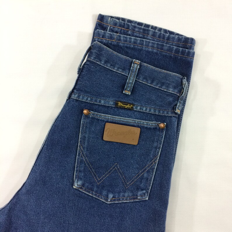 May include: Close-up of blue denim jeans with the Wrangler logo on the back pocket. The jeans have a classic five-pocket design with copper-colored rivets and a leather patch.