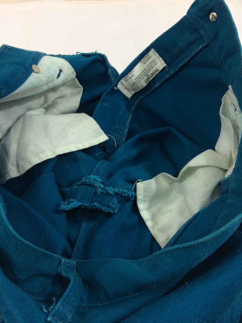 May include: Close-up of the inside of a pair of teal blue denim jeans with a white Wrangler label. The label has the text "Wrangler" and "Made in USA" printed on it.