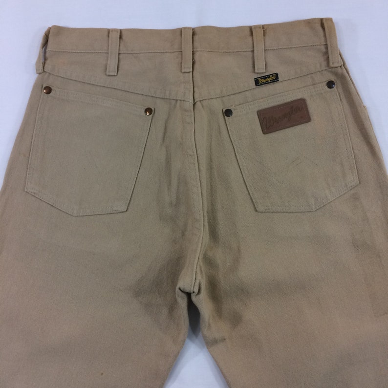 May include: A pair of tan Wrangler jeans with a brown leather patch on the back pocket that says "Wrangler".