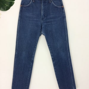 May include: A pair of blue denim jeans hanging on a wooden hanger. The jeans have a classic five-pocket design, a button and zipper closure, and a straight leg cut. The denim has a medium wash with subtle fading.