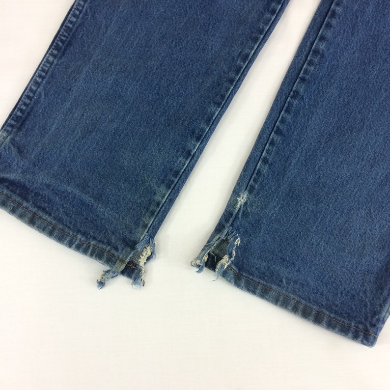 May include: Close-up of the bottom hems of a pair of blue denim jeans. The hems are frayed and distressed, showing the raw edges of the fabric.