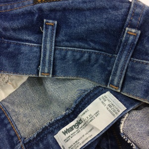 May include: A blue denim Wrangler jeans label with white text. The label reads "Machine wash hot and inside out first three times with like colors. Tumble dry. Made in Mexico of US fabric. 100% cotton. Exclusive of decoration."
